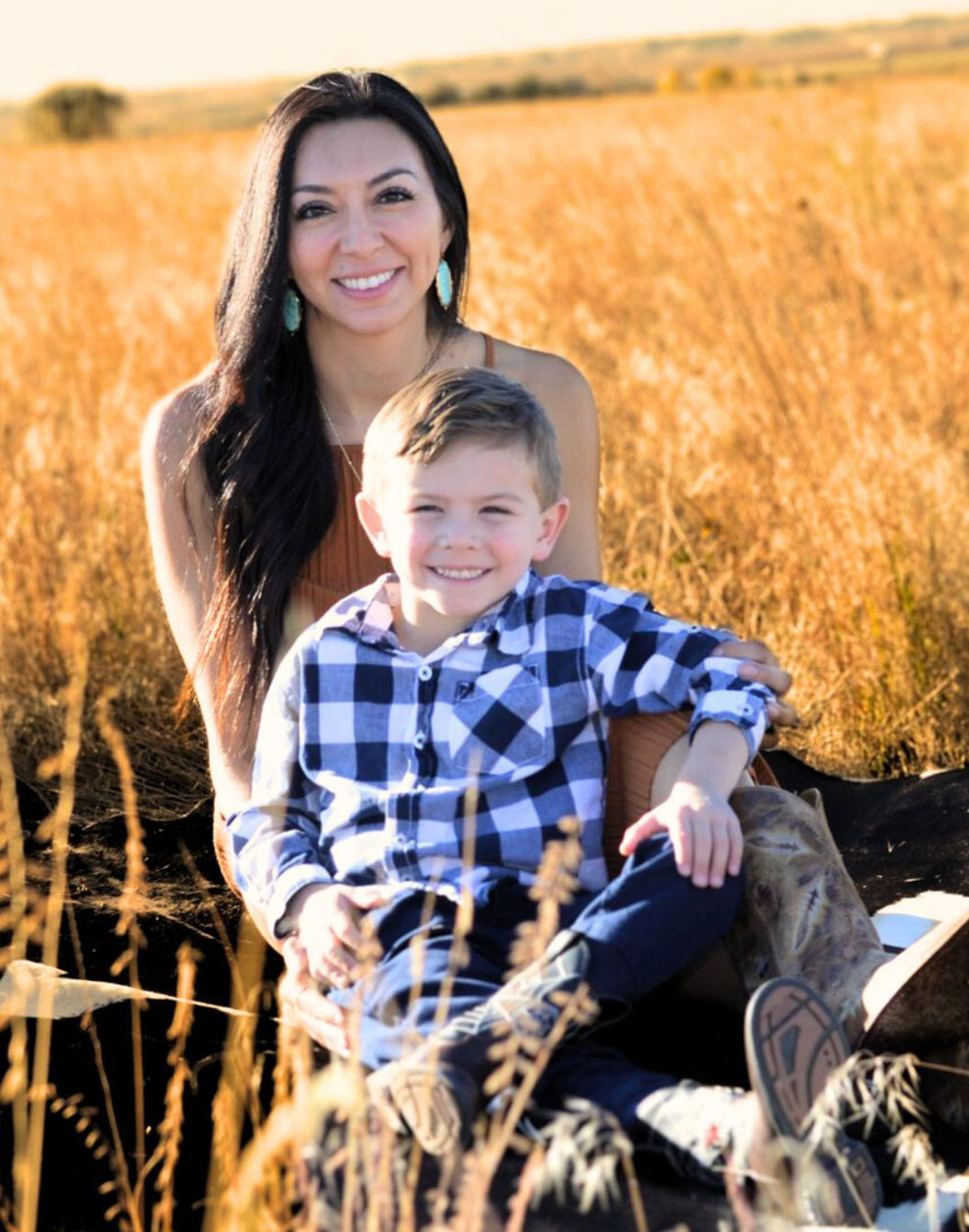 A woman and a young boy are seated on a log in a field with tall grass, smiling at the camera.