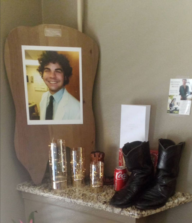 A framed photo of a smiling man wearing glasses and a blue shirt, placed on a wooden surface next to a pair of boots, a small plaque with a name, and a collection of items including a cactus, a candle, and a card.