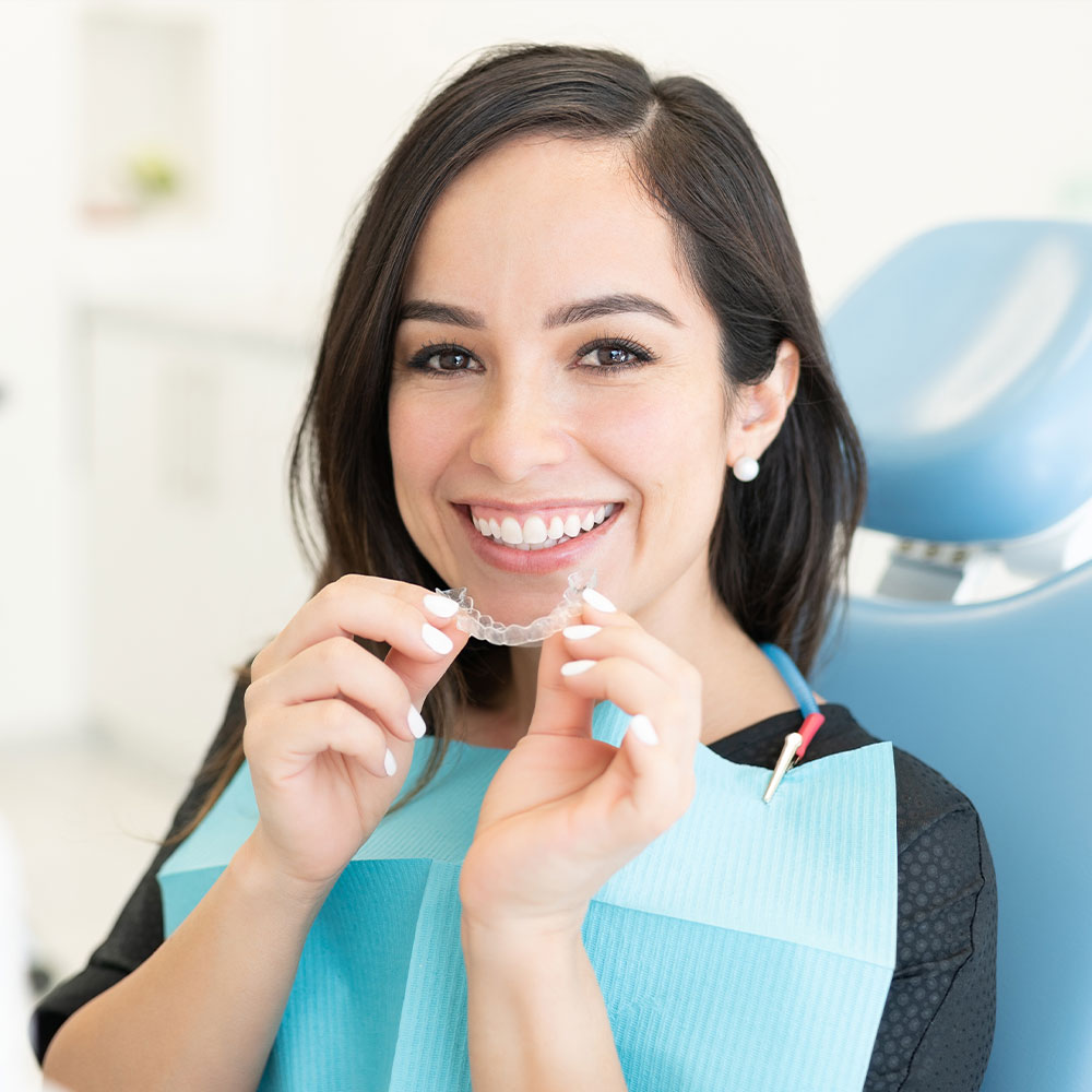 A woman wearing a dental mask and holding a toothbrush with teeth cleaning paste, smiling at the camera while seated in a dentist s chair.